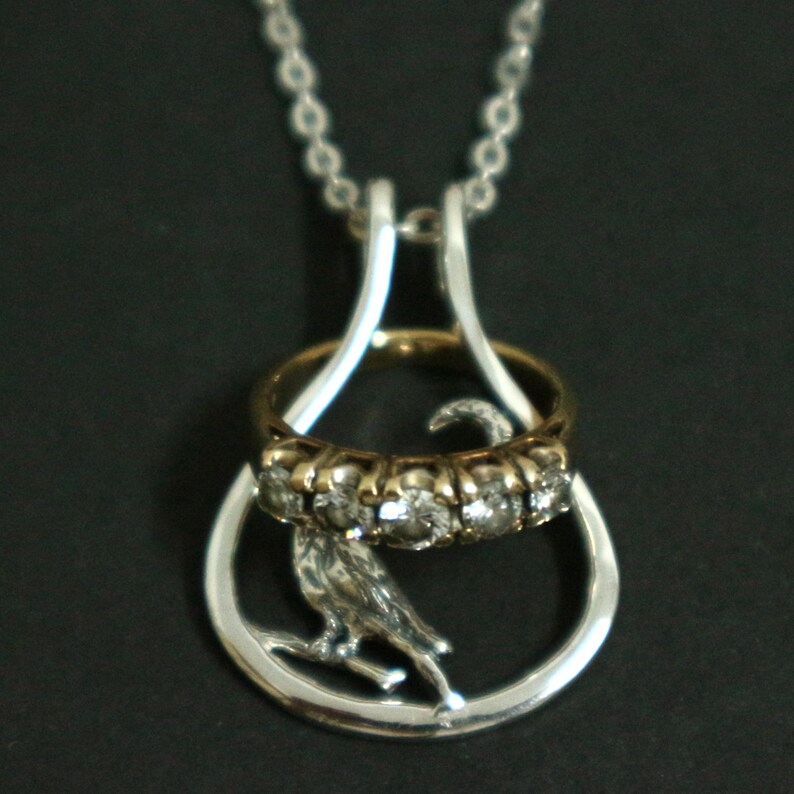 May include: Silver pendant necklace with a bird on a branch inside a teardrop frame. A gold ring with clear stones and a gold band is suspended within the frame. The necklace has a silver chain.