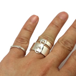 May include: Three silver rings on a finger. The top ring has a small circle charm attached to it. The middle ring is plain and the bottom ring has a small circle charm attached to it.