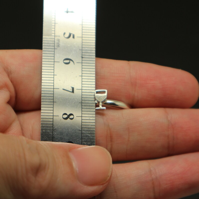 May include: A silver ring with a small wine glass charm. The ring is being measured with a ruler, showing it is approximately 7.5 mm wide.