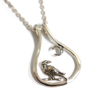 May include: Silver pendant necklace with a detailed bird perched on a branch inside a teardrop-shaped frame. A crescent moon is above the bird. The necklace has a fine chain.