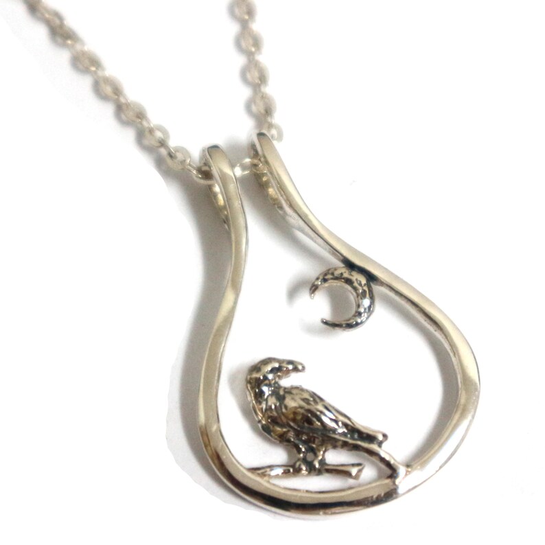 May include: Silver pendant necklace with a detailed bird perched on a branch inside a teardrop-shaped frame. A crescent moon is above the bird. The necklace has a fine chain.