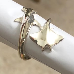 May include: A silver ring featuring a dolphin and a manta ray design. The ring wraps around the finger, with the dolphin on one side and the manta ray on the other. The ring is made of a shiny, reflective metal.