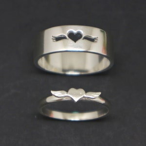 May include: Two silver rings with a heart design. The top ring has a heart with an arrow through it, while the bottom ring has a heart with wings.