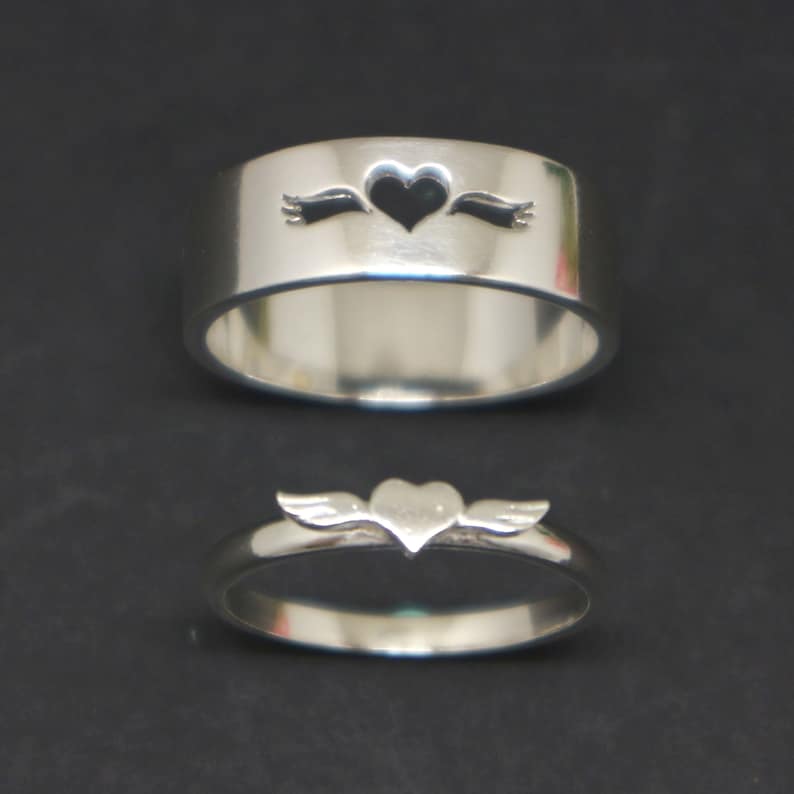 May include: Two silver rings with a heart design. The top ring has a heart with an arrow through it, while the bottom ring has a heart with wings.