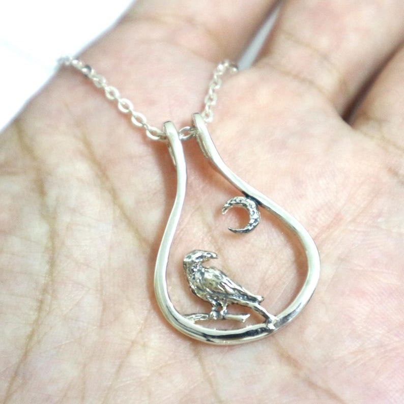 May include: Silver pendant necklace with a raven design inside a teardrop frame, with a crescent moon. The necklace is suspended from a fine chain. The raven is perched on a branch.