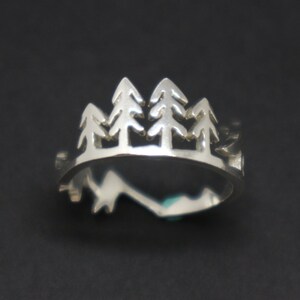 May include: A silver ring featuring a cut-out design of four pine trees. The ring is a simple band with a slightly rounded top.