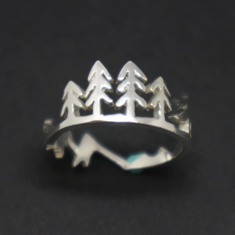 May include: A silver ring featuring a cut-out design of four pine trees. The ring is a simple band with a slightly rounded top.