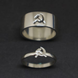May include: Two silver rings with a hammer and sickle design. The top ring is wider and has a more prominent design.