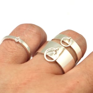 May include: Three silver rings with a simple design. The rings are stacked on a finger. The top ring has a small circle charm.