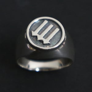 May include: A silver ring with a circular emblem featuring three downward-pointing arrows. The ring has a polished finish and a wide band. The emblem is slightly raised and has a textured appearance, set against a dark background.