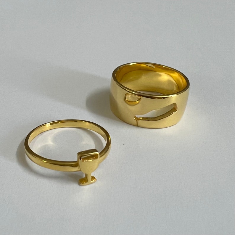 May include: Two gold rings, one with a simple band and the other with a cutout design of a wine glass. The rings are on a white surface.