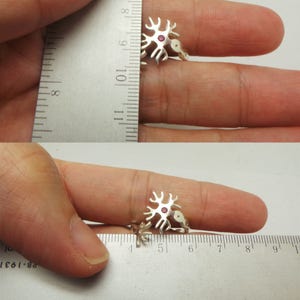 May include: A silver ring with a red gemstone in the center, shaped like a neuron. The ring is shown on a finger, with a ruler in the background.