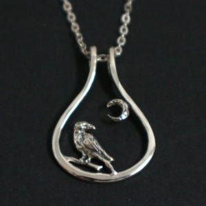 May include: Silver pendant necklace with a bird on a branch inside a teardrop frame, and a crescent moon. The pendant hangs from a fine chain. A nature-themed piece of jewellery.