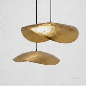May include: Two gold-coloured pendant lights suspended from black cords. The lights have an organic, abstract shape, resembling folded metal. The background is a textured white wall, creating a minimalist aesthetic.