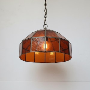 Puede incluir: Una lámpara colgante de estilo vintage con un diseño geométrico. La lámpara presenta paneles de vidrio teñido de color ámbar y un marco de metal. La lámpara cuelga de una cadena e ilumina con dos bombillas, creando un brillo cálido.