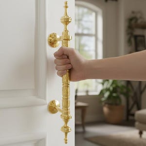 Puede incluir: Una mano abre una puerta blanca agarrando un tirador dorado con detalles ornamentados. El tirador tiene un diseño vertical con elementos decorativos. El fondo muestra un salón iluminado con luz natural.