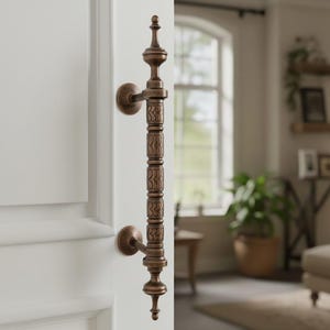 Può includere: Maniglia per porta in bronzo ornata con intagli dettagliati, montata su una porta bianca. La maniglia ha un design verticale con elementi decorativi e una parte superiore a punta. Lo sfondo mostra un soggiorno.