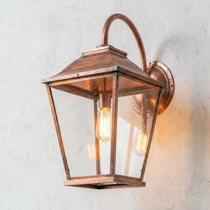 May include: Copper-colored outdoor wall lantern with a clear glass enclosure and a curved arm. The lantern features a vintage-style light bulb and is mounted on a textured white wall. The fixture has a classic, elegant design.