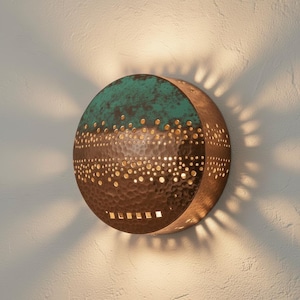 May include: A circular wall sconce in copper with a teal top, featuring a textured finish and numerous perforations. The perforations allow light to shine through, creating a starburst effect on the wall. The sconce is mounted on a white wall.