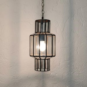 May include: A hanging pendant light fixture with a geometric design. The light has a black metal frame with clear glass panels. A single light bulb is visible inside the fixture. The light hangs from a chain.