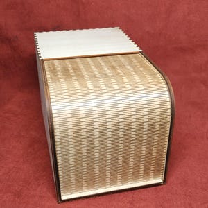 May include: A wooden box with a curved, textured front and a flat top. The box is made of light-colored wood with a repeating pattern of thin lines. The top is a lighter shade of wood. The box is on a red background.
