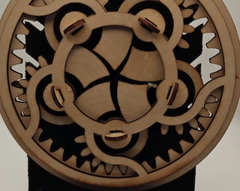 Könnte beinhalten: Ein kreisförmiges mechanisches Holzpuzzle mit einem komplizierten Getriebesystem aus ineinandergreifenden Zähnen und einer zentralen rotierenden Komponente. Das helle Holz kontrastiert mit einer dunklen Innenschicht und zeigt ein komplexes Zahnradsystem.