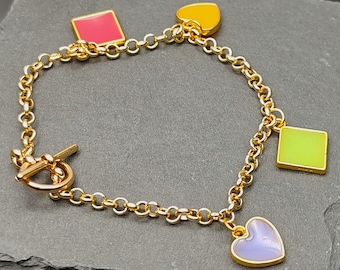 Könnte beinhalten: Ein goldfarbenes Gliederkettarmband mit Knebelverschluss und vier Emaille-Anhängern in verschiedenen Formen und Farben, darunter ein rotes Quadrat, ein gelbes Herz, ein lindgrünes Quadrat und ein hellviolettes Herz, die an den Gliedern hängen.