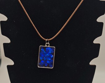 Könnte beinhalten: Eine rechteckige Anhängerkette mit einem silberfarbenen Metallrahmen, der mit tiefblauen, kreisförmigen Glitzerpartikeln gefüllt ist. Der Anhänger hängt an einer dünnen, braunen Kordel, die um die Oberseite des Ständers verläuft.
