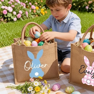 Peut inclure: Deux sacs fourre-tout de Pâques en toile de jute avec des poignées, remplis d'œufs de Pâques colorés. Un sac a un motif de lapin bleu et le nom "Oliver", l'autre a un lapin à carreaux roses et le nom "Evely". Un enfant place des œufs dans les sacs.