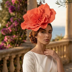 Könnte beinhalten: Ein großer, leuchtend korallenoranger floraler Kopfschmuck mit geschichteten, strukturierten Blütenblättern, die einer blühenden Blume nachempfunden sind. Das Accessoire ist im Haar befestigt und zeigt eine detaillierte Mitte mit feinen Staubgefäßen.