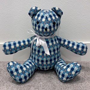 May include: A handmade teddy bear with a blue and white patterned fabric. The bear has a white ribbon tied around its neck and black button eyes. The bear is sitting on a gray carpet.