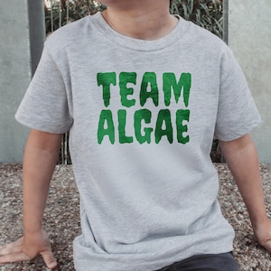 Peut inclure: Un t-shirt gris chiné à manches courtes avec le texte "TEAM ALGAE" imprimé en lettres vertes grasses et dégoulinantes sur la poitrine. Le vêtement présente une encolure ronde classique et une coupe standard avec un tissu texturé.
