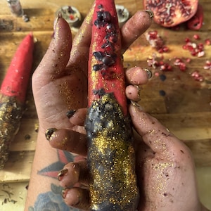 May include: A red candle with black accents and gold glitter is held in a hand. The candle is decorated with dark spots and gold glitter. A second candle is visible in the background. A halved pomegranate is also visible.