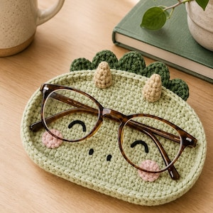 May include: A light green oval crochet tray featuring a dinosaur-inspired design with dark green spikes, two small beige horns, embroidered black eyes, and pink cheeks. A pair of brown tortoiseshell eyeglasses rests on top of the crochet tray.