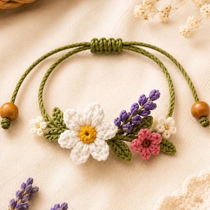 Op de afbeelding: Een handgemaakte verstelbare armband met een groen koord, een schuifknoopsluiting en twee houten kralen. Het middendeel is versierd met gehaakte bloemenelementen, waaronder een grote witte margriet, paarse lavendeltakjes en een klein roze bloemetje.