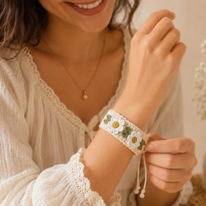 May include: A cream-colored crochet bracelet featuring a repeating pattern of white daisy flowers with yellow centers and small green leaves. The bracelet has a thin adjustable drawstring closure with two small knotted ends hanging from the side.