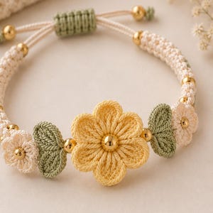 May include: A macrame bracelet featuring a central yellow flower, flanked by smaller cream-colored flowers and sage green leaves. The design incorporates polished gold-tone metal beads between the floral elements and includes an adjustable cord closure.