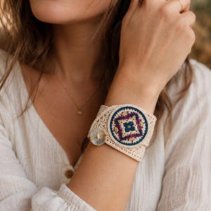 May include: A wide beige crochet cuff bracelet featuring a central circular medallion with intricate geometric embroidery in shades of deep purple, teal, and gold. The bracelet is secured with a single round, mottled grey button closure.