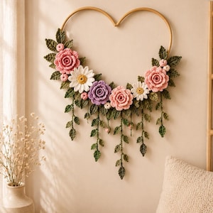 May include: A heart-shaped wall hanging featuring crocheted flowers and leaves. The arrangement includes pink and purple roses, white daisies with yellow centers, and green foliage. Several crocheted vines with small leaves hang downward from the frame.