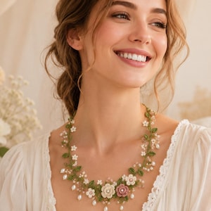 Puede incluir: Un delicado collar floral con intrincadas enredaderas verdes de ganchillo, pequeñas flores de ganchillo blancas y rosas, y cuentas blancas colgantes similares a perlas. La pieza está hecha con hilo fino y decorada con diminutas cuentas transparentes.