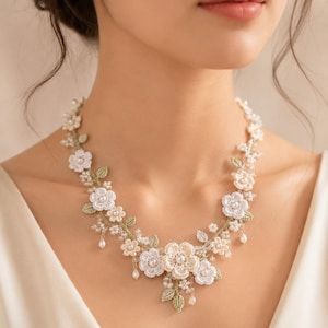Può includere: Una collana floreale con intricati fiori all'uncinetto bianchi e color crema, piccole foglie verdi all'uncinetto e delicate perle bianche pendenti. Il design botanico è disposto in un motivo simmetrico attorno al collo con dettagli in filo.