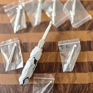 Könnte beinhalten: Eine Sammlung von Häkelnadeln und Werkzeugen. Ein Haken hat einen weiß-schwarzen marmorierten Griff. Mehrere Haken sind in kleinen, durchsichtigen Plastiktüten verpackt. Die Haken sind silberfarben und liegen auf einer Holzoberfläche.