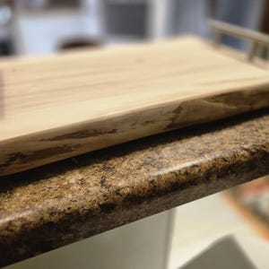 May include: A rectangular wooden serving tray with natural wood grain and a live edge. It has two brushed metal handles. The light brown tray is likely handmade, suitable for serving food or as a decorative item. The tray measures approximately 30 cm in length.