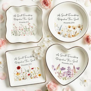 Puede incluir: Cuatro platos de cerámica blanca en formas cuadradas, redondas y de corazón con bordes dorados. Cada plato presenta una ilustración de flores silvestres coloridas y el texto negro A Sweet Friendship Refreshes The Soul Proverbs 27:9.