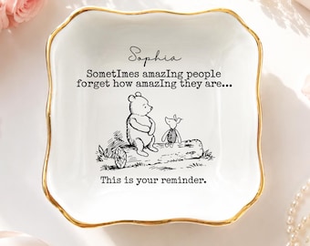 Può includere: Un piattino quadrato in ceramica bianca con un bordo smerlato dipinto in oro. Il centro presenta un'illustrazione a inchiostro nero di Winnie the Pooh e Pimpi seduti su un tronco, con il testo: Sophia, SomeTimes amazIng people forget how amazIng they are... This is your reminder.