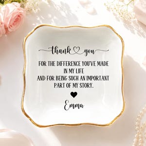 May include: A square white ceramic trinket dish with scalloped edges and a gold-painted rim. The centre features black text that reads: thank you FOR THE DIFFERENCE YOU'VE MADE IN MY LIFE AND FOR BEING SUCH AN IMPORTANT PART OF MY STORY. Emma.