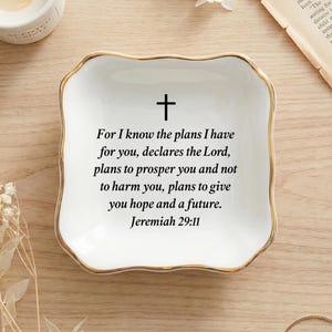 May include: White ceramic trinket dish with a gold rim and a black cross. The dish features the text "For I know the plans I have for you, declares the Lord...Jeremiah 29:11". Decorative item for jewelry or small items.