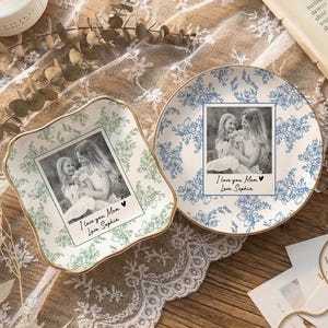 Puede incluir: Una taza de cerámica blanca con asa negra y el texto "But First, Coffee" en negrita negra. La taza se encuentra sobre un fondo borroso, sugiriendo un ambiente acogedor.