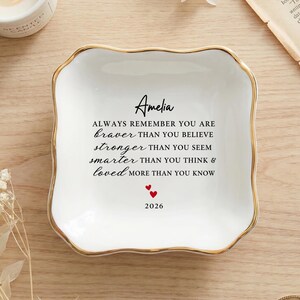 Peut inclure: Un vide-poche carré en céramique blanche avec des bords festonnés et un bord doré. Le centre présente un texte noir indiquant Amelia, Always remember you are braver than you believe, stronger than you seem, smarter than you think & loved more than you know, avec deux petits cœurs rouges et l'année 2026 imprimés en dessous.