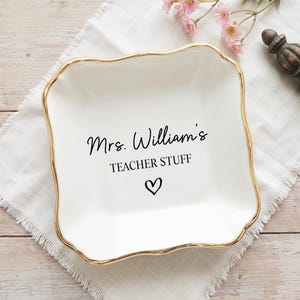 Może przedstawiać: Biały ceramiczny talerz ze złotą obwódką, z napisem "Mrs. William's TEACHER STUFF" i symbolem serca. Talerz leży na białej tkaninie, z drewnianym elementem dekoracyjnym i różowymi kwiatami w pobliżu.
